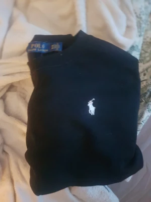 Svart tröja från Polo Ralph Lauren - Klassisk svart tröja från Polo Ralph Lauren med rund halsringning och den ikoniska vita broderade loggan på bröstet. Tröjan har en enkel och stilren design som passar till det mesta. Perfekt för dig som gillar tidlös stil.