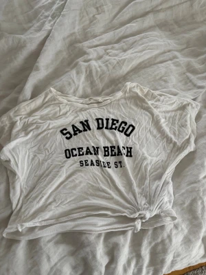 Vit croppad t-shirt med tryck - Säljer en vit croppad t-shirt med svart text framtill. T-shirten har korta ärmar och en knytning i sidan för en avslappnad look. Perfekt till sommaren och enkel att matcha med jeans eller shorts. Extra snyggt med ett linne under🤩💓💓🤍