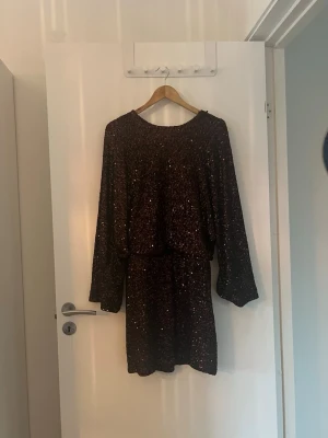 Svart glittrig kortklänning - Säljer en svart kortklänning med glittriga detaljer. Klänningen har långa ärmar och en lös passform i midjan. Perfekt för dig som vill sticka ut med lite extra glitter. Ingen synlig logga eller märke.