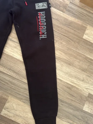 Svarta Hoodrich mjukisbyxor - Svarta mjukisbyxor från Hoodrich med tryckt logga i vitt och rött längs ena benet. Byxorna har ribbade muddar vid bensluten och dragsko i midjan med röda detaljer. Perfekta för en avslappnad streetwear-look.