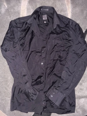 Svart skjorta från Calvin Klein - Snygg svart skjorta från Calvin Klein med klassisk krage och knäppning framtill. Skjortan har långa ärmar med manschettknappar och en diskret bröstficka. Perfekt för en stilren look och tillverkad i ett mjukt tyg.
