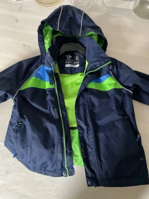 Neo Mon Do Mörkblå vindjacka från Neomoon - Sportig mörkblå vindjacka från Neomoon med gröna och blå detaljer. Jackan har huva, dragkedja och meshfoder i neonfärg. Perfekt för blåsiga dagar och har en cool, ungdomlig stil med kontrastfärger och praktiska fickor.