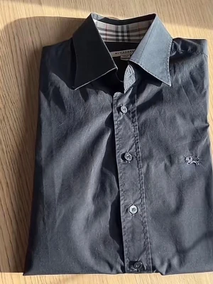 Svart skjorta från Burberry - Klassisk svart skjorta från Burberry med knappar framtill och ikoniskt rutigt mönster på insidan av kragen. Skjortan har lång ärm och broderad logga på bröstet. Perfekt för en stilren look. Pris kan diskuteras