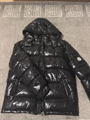 Svart glansig dunjacka från Moncler - Säljer en svart, glansig dunjacka från Moncler med huva och dragkedja framtill. Jackan har två snedställda fickor med dragkedja och Moncler-logga på ärmen. Perfekt för kalla vinterdagar och riktigt snygg streetstil.