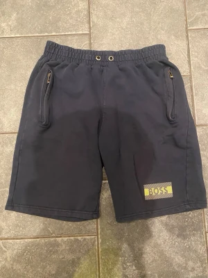 Mörkblå BOSS shorts med dragkedjor - Snygga mörkblå shorts från BOSS med gul logga på vänster ben. Shortsen har elastisk midja med snörningshål och två fickor med dragkedja. Perfekta för chill dagar eller när du vill ha en clean look.