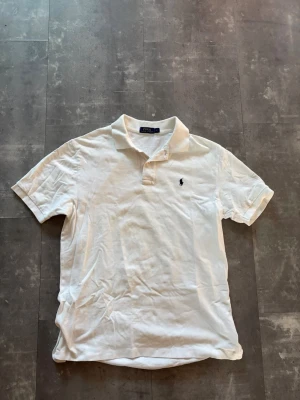 Vit pikétröja från Polo Ralph Lauren - Klassisk vit pikétröja från Polo Ralph Lauren med korta ärmar och broderad logga på bröstet. Tröjan har krage och knäppning framtill. Perfekt för en clean och stilren look.
