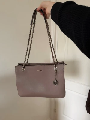 Taupe handväska från DKNY - Snygg taupefärgad handväska från DKNY med guld kedjehandtag och läderdetaljer. Väskan har en stilren, rektangulär form och ett dekorativt DKNY-hänge framtill. Perfekt för dig som gillar klassisk och trendig design. Man får plats med en dator, iPad osv. 