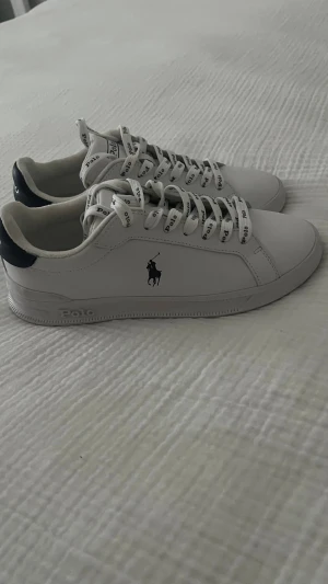 Vita Polo Ralph Lauren sneakers - Stilrena vita sneakers från Polo Ralph Lauren med svart logga på sidan och svarta detaljer vid hälen. Skorna har vita snören med Polo-tryck och en klassisk låg siluett. Tillverkade i slätt läder för en clean och modern look. Tillkommer helt vita skosnören dessa är aldrig använda. Jag har användent skorna typ två gånger. 