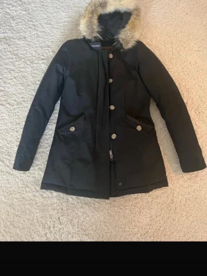 Marinblå parkas från Woolrich med äkta päls - Snygg marinblå parkas från Woolrich med huva och äkta päls. Jackan har knappar framtill, två stora fickor med lock och är fodrad för extra värme. Perfekt för kalla dagar och har en klassisk, rak passform.