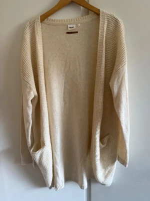 Beige lång kofta från Object - Lång, öppen kofta i beige från Object. Koftan har två fickor framtill, ribbade muddar och är stickad i ett mjukt material med struktur på ärmar och kanter. Perfekt för lager på lager och enkel att matcha med olika outfits.