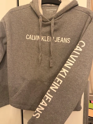 Grå hoodie från Calvin Klein Jeans - Snygg grå hoodie från Calvin Klein Jeans med vit logga tryckt både på bröstet och längs ena ärmen. Hoodien har en klassisk huva med dragsko och en stor magficka. Mjuk insida för extra komfort, perfekt för en avslappnad stil.