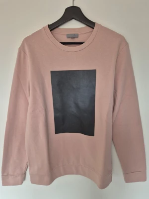 Rosa sweatshirt från COS med tryck - En stilren rosa sweatshirt från COS med ett svart fyrkantigt tryck på bröstet. Tröjan har rund halsringning och långa ärmar. Perfekt för en avslappnad och trendig look.