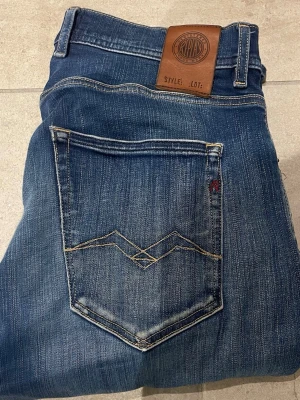 Blå slitna jeans från Replay - Säljer ett par blå jeans från Replay med snygg slitning på ena benet och klassiska kontrastsömmar. Jeansen har fem fickor, normal midja och en patch i läder baktill. Perfekta för en avslappnad och trendig look. 31/32
