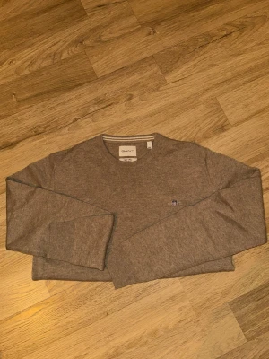 Beige stickad tröja från GANT - Snygg beige stickad tröja från GANT med rund halsringning och diskret broderad logga på bröstet. Tröjan har långa ärmar och är tillverkad i ett mjukt, tunt material som passar perfekt för lager på lager. Klassisk och stilren design.