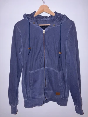 Blå hoodie från Woolrich med dragkedja - Säljer en blå hoodie från Woolrich i storlek M. Tröjan har huva med snörning, dragkedja framtill och två stora fickor. Tillverkad i mjuk bomull och har ribbade muddar vid ärmslut och nederkant. Perfekt för chill dagar eller när du vill ha en avslappnad stil. Passar S-M
