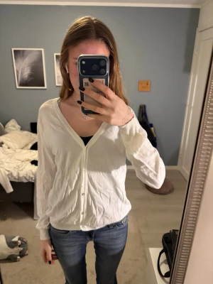 Vit långärmad kofta med knappar - Säljer en enkel vit kofta med v-ringning och knäppning framtill. Koftan har långa ärmar och är tillverkad i ett tunt, mjukt material som passar perfekt att ha över ett linne eller t-shirt.