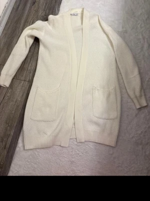 Vit lång cardigan med fickor - Säljer en lång, vit cardiganjacka med två stora fickor framtill. Jackan är stickad med ribbade muddar vid ärmslut och nederkant. Perfekt att slänga över en t-shirt för en chill look.