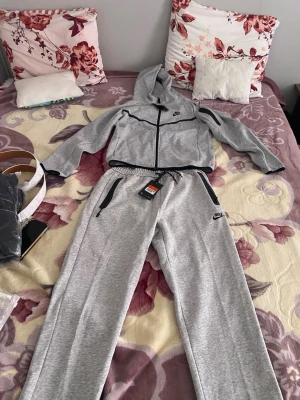 Gråa Nike Tech Fleece hoodie och byxor  - Snygga gråa Nike Tech Fleece mjukisbyxor och hoodie med svarta dragkedjor och logga på benet. Byxorna har smal passform och är tillverkade i mjukt och stretchigt material som ger en sportig look. Perfekta för chill eller träning.