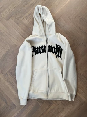 Vit zip hoodie med svart tryck - Säljer en vit hoodie med dragkedja och stor huva. På bröstet finns ett fett svart tryck i gotisk stil. Hoodien har två fickor fram och ribbade muddar vid ärmar och nederkant. Perfekt för dig som gillar streetwear vibes.