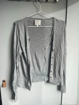 Ljusgrå tunn kofta med knappar - Ljusgrå tunnstickad kofta med långa ärmar och knäppning framtill. Klassisk rak passform och ribbade muddar vid ärmslut och nederkant. Perfekt att slänga över en t-shirt för en enkel och clean look.