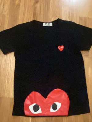 Cdg t shitt - Svart t-shirt från Comme des Garçons PLAY med ikoniskt rött hjärta med ögon på bröstet och ett stort hjärta med ögon tryckt längst ner. Klassisk passform och rund hals. Perfekt för dig som gillar streetwear och vill sticka ut. Passar som xs och s