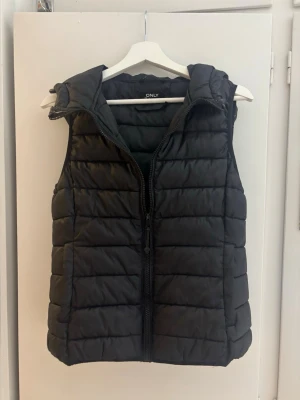 Svart dunväst med huva - Snygg svart dunväst från ONLY med quiltad design och dragkedja framtill. Västen har en praktisk huva och är perfekt för lager-på-lager-stil. Tillverkad i ett lätt och värmande material som håller dig varm under kyliga dagar. 