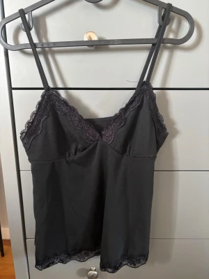 Svart linnetopp med spetsdetaljer - Stilren svart linnetopp med tunna axelband och v-ringad spetskant både upptill och nedtill. Tillverkad i mjukt material som känns skönt mot huden. Perfekt att matcha med jeans eller kjol för en trendig look.