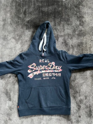 Mörkblå hoodie från Superdry - Mörkblå hoodie från Superdry med stor huva och vita dragsnören. Framsidan har ett blommigt Superdry-tryck och text i vitt och rosa. Klassisk känguruficka framtill och ribbade muddar. Perfekt för en avslappnad och trendig stil.