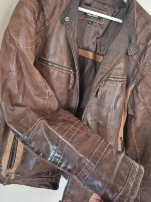 Brun skinnjacka från KALLTAN - Snygg brun vintage skinnjacka från Molltan med dragkedjor på bröstet och sidorna. Jackan har coola sömdetaljer, ståkrage med tryckknapp och kontrasterande paneler i ljusare brunt. Perfekt för både vår, svalare sommar och höst. Har några slitningar.