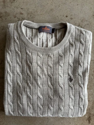 Beige  kabelstickad tröja Morris - Kabelstickad merinoulls tröja från Morris i beige med rund halsringning och ribbade muddar. Tröjan har ett diskret Morris-emblem broderat på bröstet och är tillverkad i ett mjukt material som passar perfekt till lager på lager.