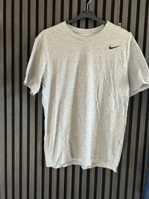 Grå Nike t-shirt - Snygg grå t-shirt från Nike med klassisk svart logga på bröstet. Tillverkad i mjuk bomull och har en enkel, normal passform med rund hals och korta ärmar. Perfekt till vardags eller träning.