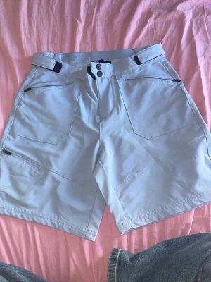 Ljusblåa outdoor  shorts med fickor - Säljer ett par ljusblåa shorts med stora framfickor och knappstängning. Shortsen har en rak passform och bälteshällor i midjan. Perfekta för varma dagar och enkel att matcha med olika toppar.Funktionsmaterial