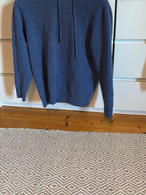 Blå hoodie i mjukt material - En stilren blå hoodie med huva och dragsnören. Tillverkad i ett mjukt, stickat material som känns skönt mot huden. Hoodien har långa ärmar och ribbade muddar vid ärmslut och nederkant. Perfekt för en avslappnad look.
