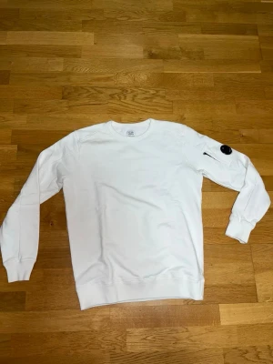 Vit sweatshirt från C.P. Company - Vit sweatshirt från C.P. Company med rund halsringning och lång ärm. Tröjan har en ikonisk svart linsdetalj på vänster ärm och en liten dragkedjeficka. Perfekt för en clean och stilren look.