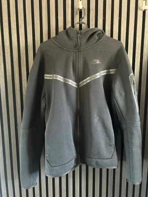 Svart Nike tech flecce - Snygg svart Nike tech flecce från Nike med hel dragkedja framtill och reflekterande detaljer över bröstet. Tröjan har huva och en ficka med dragkedja på ena ärmen. Perfekt för dig som gillar sportig och clean stil.