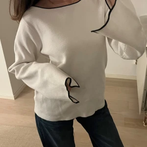 Vit tröja med kontrastsömmar - Säljer en vit långärmad tröja med svarta kontrastsömmar längs halsringning och ärmslut. Tröjan har en avslappnad passform och ett unikt broderat motiv på ena ärmen. Perfekt för dig som gillar minimalistisk stil med en twist.