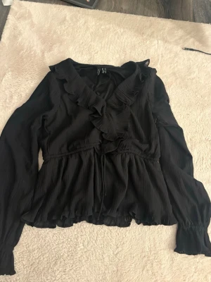 Svart volangblus med knytband - Snygg svart blus med volangdetaljer längs ringningen och knytband i midjan. Blusen har långa ärmar och lätt rynkad nederkant för en feminin look. Tillverkad i ett tunt, strukturerat tyg som ger en luftig känsla.