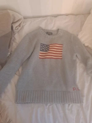 Ljusgrå stickad tröja Ralph Lauren - Ljusgrå stickad tröja från Ralph Lauren med amerikansk flagga på bröstet och RL-broderi nere vid fållen. Tröjan har rund halsringning, långa ärmar och ribbstickade muddar. Perfekt för dig som gillar klassisk och avslappnad stil.