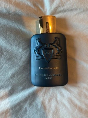 Layton Exclusif Parfums de Marly - Lyxig parfymflaska i matt svart med guldfärgat lock från Parfums de Marly. Flaskan har upphöjt emblem med två hästar och texten 'Layton Exclusif'. Kommer i originalförpackning med 'Edition Royale' och rymmer 125 ml.