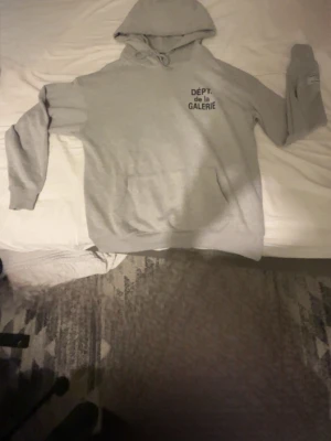 Grå hoodie från Gallery Dept. - Snygg grå hoodie från Gallery Dept. med trycket 'DÉPT de la GALERIE' på bröstet. Klassisk känguruficka framtill och justerbar huva med dragsko. Perfekt för en avslappnad och trendig streetwear-look.