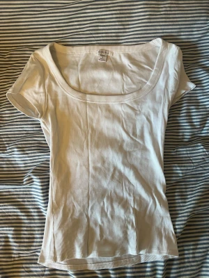 Vit ribbad topp från Amsu - Snygg vit ribbad topp från Amsu med korta ärmar och bred, rundad urringning. Tillverkad i mjuk bomullsmix som sitter skönt och har en slim passform. Perfekt att styla med jeans eller kjol för en enkel och clean look.
