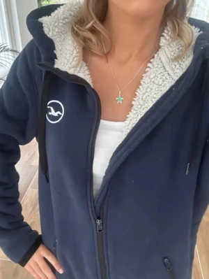 Marinblå hoodie från Hollister - Mysig marinblå hoodie från Hollister med vit logga på bröstet. Insidan är fodrad med fluffigt, vitt teddyfoder som gör den extra varm. Hoodien har dragkedja, två fickor och justerbar huva med vita snören. Perfekt för chill dagar och sköna outfits.