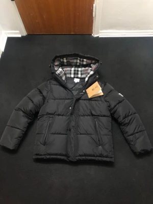 Svart pufferjacka från Burberry - Svart pufferjacka från Burberry med avtagbara ärmar, så du kan bära den som väst. Jackan har huva med rutigt foder i svart, vitt och rött, samt två dragkedjefickor fram. Burberry-logga på vänster ärm och snygga detaljer med knappar och dragkedja.