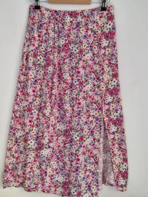 Blommig midikjol med slits - Säljer en midikjol med blommigt mönster i rosa, lila, gul och vitt. Kjolen har en snygg slits på sidan och är perfekt för dig som gillar färgglada och somriga plagg. Materialet känns lätt och flowy, vilket gör den skön att bära under varma dagar.