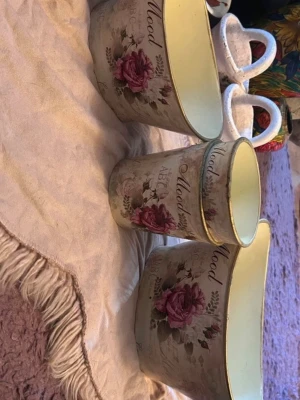 Blommiga krukor med text Mood - Säljer tre dekorativa krukor i metall med vintageinspirerat blommönster i rosa och grönt samt texten 'Mood' på framsidan. Krukorna har en ljusbeige bakgrund och guldfärgade kanter. Perfekta som inredningsdetaljer eller för växter.