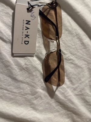 Solglasögon - Snygga solglasögon från NA-KD med rektangulära, bruna linser och tunna guldfärgade bågar. Glasögonen har minimalistisk design och svarta detaljer på skalmarna.