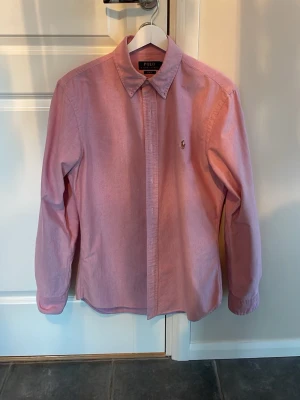 Rosa skjorta från Polo Ralph Lauren - Klassisk rosa skjorta från Polo Ralph Lauren med lång ärm och knappar framtill. Skjortan har en broderad logga på bröstet och är tillverkad i mjuk bomull. Perfekt för en clean och stilren look.
