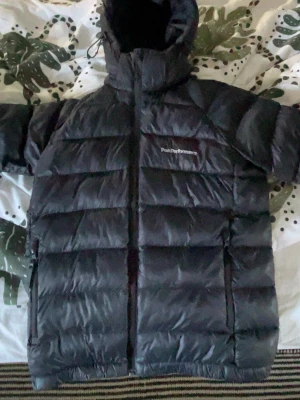 Svart dunjacka från Peak Performance - Säljer en svart dunjacka från Peak Performance med huva och dragkedja framtill. Jackan har en puffig design med breda kanaler och logga på bröstet. Perfekt för kalla vinterdagar och riktigt varm tack vare dunfyllningen.