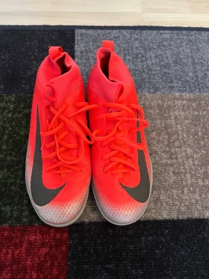 Nike röda fotbollsskor med högt skaft - Säljer ett par grymma fotbollsskor från Nike i en riktigt snygg röd färg med svart swoosh på sidan. Skorna har högt skaft för extra stöd och är tillverkade i syntetmaterial med snörning. Perfekta för dig som vill sticka ut på planen!