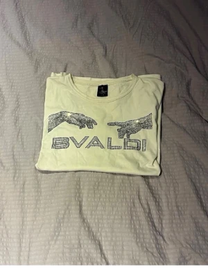 BVALDI T-shirt  - Vit t-shirt från Bvaldi med glittriga strasstenar som formar två händer och texten 'BVALDI' på bröstet. Klassisk rund halsringning och korta ärmar. Perfekt för dig som gillar unika detaljer och lite extra bling.